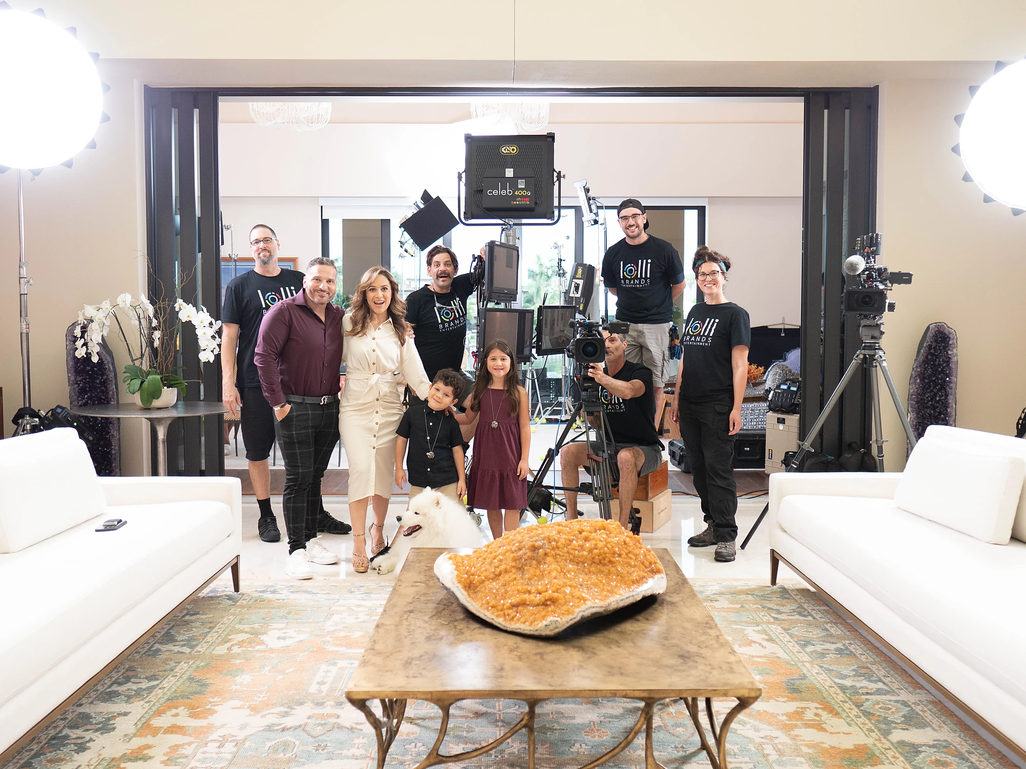Crew in Lolli Brands Entertainment shirts with a couple and two children on a living-room set: Celeb 400 light, monitor stack, cinema cameras, amethyst geode