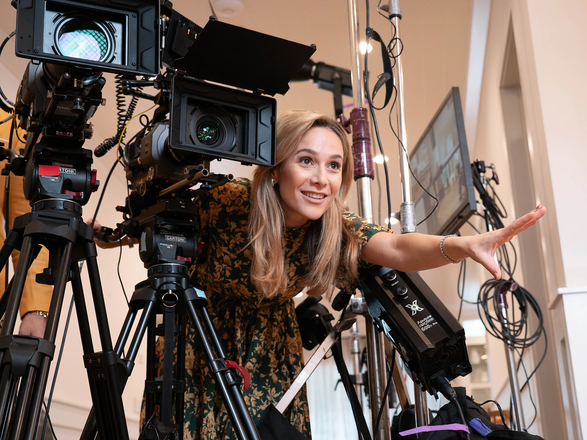 Woman leaning in beside a cinema camera on sticks with her arm extended to frame a shot, cables and light stands behind